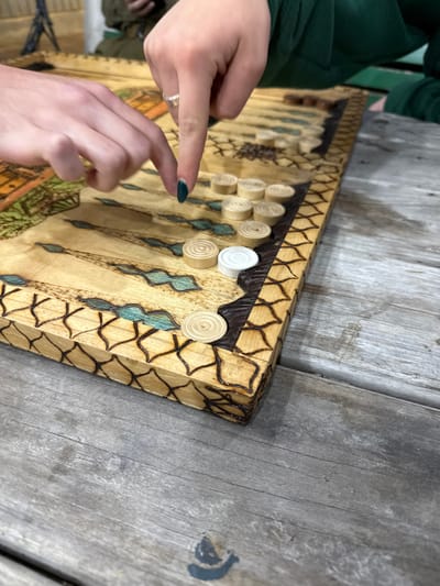 The image shows a wooden backgammon board with light brown, white, and dark brown pieces. The board is sitting on a wooden table. Two hands are reaching towards the board, with one hand in the foreground placing a piece. The image was likely taken in Mazkeret Batya, Israel. The coordinates provided place the location in a residential area near a park. It is possible that the image was taken in a private home, a park, or a public area.