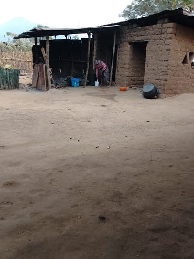 A rural scene shows a woman in a pink and floral robe filling a bucket near a weathered, makeshift structure. This structure is a combination of corrugated metal and clay brick, with a covered open area next to a small, enclosed room. Several buckets and containers are scattered around the building. The ground is bare dirt, and a simple wooden fence stands in the background.