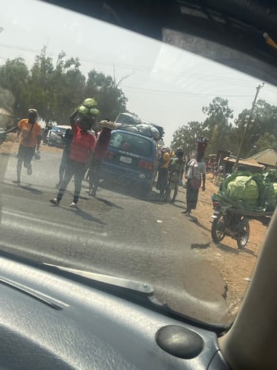 The image captures a busy street scene in Hoss, Nigeria, viewed from within a vehicle under a bright, hazy sky. In the mid-ground, a person wearing a red shirt and dark pants walks left-to-right, balancing four large green cabbages on their head. Behind them, another individual carries a tray of eggs on their head. To the left, a person in an orange shirt and dark pants moves rapidly from left to right. A blue van, bearing the text "ISLAMIC PRAYERS" on its rear window and a "GH" country sticker, is heavily loaded with bundled goods on its roof rack; its license plate partially reads "IKJ 1". To the right of the van, several individuals are present; one person in a light-colored top carries a red container on their head and a bag over their shoulder, walking right-to-left. Further right, a motorcycle is stationary or moving slowly, laden with a substantial quantity of green cabbages on a rear-mounted flatbed. The scene features a paved road, a dirt shoulder, distant trees with green foliage, a utility pole, and partially visible light-colored buildings in the background.