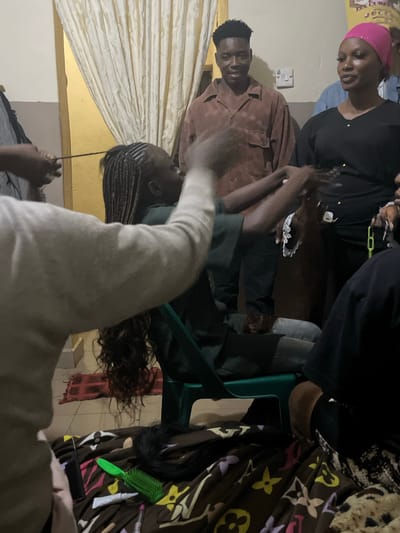 The image depicts an indoor scene, possibly a home or salon, in Durumi, Nigeria. A young woman is seated on a green chair, with her hair being braided by at least two individuals whose hands are visible. The braids are long and extend down her back. Scattered around the floor, on what appears to be a patterned blanket, are various hair care items, including a green brush, a tube of what looks like hair product, and a black comb. In the background, a young man stands wearing a brown button-up shirt, and a woman in a pink headscarf and black top is also present. The environment appears to be dimly lit with light-colored curtains in the background.