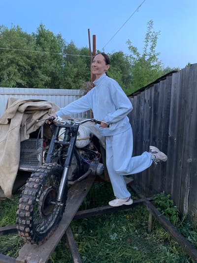 The image shows a person lying on top of a dismantled motorcycle in Doma 45 Kilometr, Russia. The individual is wearing a light blue tracksuit and sneakers, smiling and looking towards the camera. The motorcycle appears to be undergoing repairs or is stored in a disassembled state, partially covered with a tan fabric. The background includes a wooden fence, vegetation, and sections of buildings. The scene suggests an outdoor setting within the city of Doma 45 Kilometr.