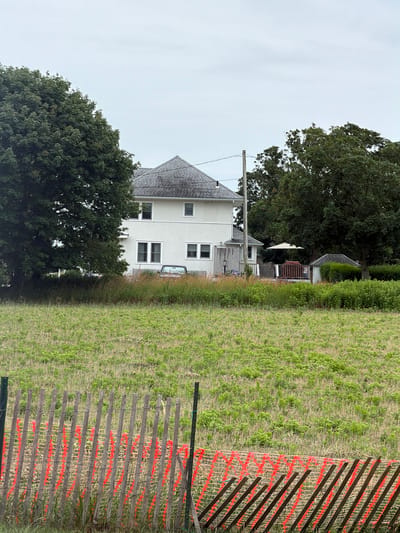 The media file displays an outdoor scene, likely in Mattituck, USA, on an overcast day. The primary subjects include a two-story house with a grey shingled roof and light-colored siding, featuring multiple windows. Adjacent to the house are mature trees, one of which is prominent in the foreground, with dense green foliage. To the right of the house, a grassy field stretches towards a fence. This fence is constructed of wooden slats and is partially covered with orange mesh netting, giving it a striped appearance.
In the background, behind the main house, a smaller structure with a peaked roof and a wooden deck is visible, partially obscured by trees and bushes. A white umbrella can be seen on the deck. A light-colored car is parked near the house, facing away from the viewer.
The scene appears to be a residential property, possibly near a rural or semi-rural area given the expanse of the field. The overcast sky suggests it is either early morning, late afternoon, or a generally cloudy day, and the overall mood is calm and uneventful. There is no discernible activity taking place. No visible text is present in the image.