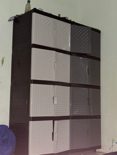 A tall, multi-tiered plastic cabinet stands in a corner against a light green wall in Tupah, Indonesia. The cabinet is dark brown on the left and alternates between light gray and dark gray on the right, with each tier having two doors. The light gray doors have a raised geometric pattern, while the dark gray doors have a darker, embossed geometric pattern. The top of the cabinet holds a clear plastic bottle with a green cap and a yellow label, along with other unidentifiable items. Below the cabinet, a folded blue cloth is visible on the floor. The wall shows signs of wear, with patches of peeling paint.