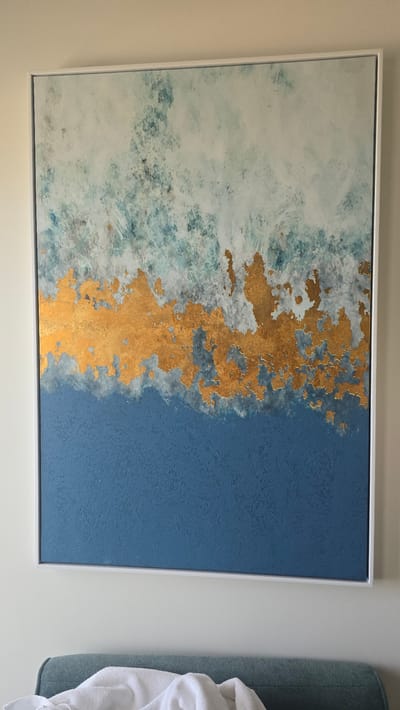 The image displays a framed abstract painting with a white border. The painting is divided into two main sections: a left side with swirling patterns of white, light blue, and hints of green, and a right side of solid, textured deep blue. A prominent, irregular strip of textured gold leaf runs vertically down the center, separating the two color fields and creating a striking contrast. The textured nature of the gold leaf suggests a tactile element to the artwork.
The painting is hung on a light-colored wall. To the right of the painting, a portion of a teal-colored upholstered object, possibly a chair or sofa, is visible. A crumpled white cloth, resembling a towel or blanket, is draped over this object. The lighting appears to be natural, casting soft shadows and highlighting the textures of the artwork. There are no visible people, specific activities, or discernible text in the image. The overall impression is one of modern decor with an emphasis on color and texture. The setting is domestic, likely within a home.