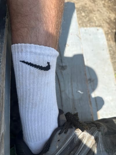 A close-up, overhead shot shows a person's left leg and foot, wearing a white Nike sock with a black Nike logo and a gray athletic shoe. The leg is hairy, and the sock is pulled up to mid-calf. The shoe laces are dark and textured. The background is a wooden surface, possibly a bench or deck, in an outdoor setting. Sunlight casts shadows across the scene. The overall impression is casual and outdoor-oriented.