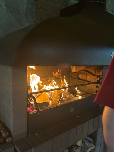 An indoor fireplace is filled with burning logs. The fireplace is constructed of light-colored bricks and has a dark metal hood. A person wearing a red shirt is partially visible on the right side of the image. Wood is stacked in the lower portion of the fireplace structure. The scene appears to be from a home in Ryabinino, Russia.