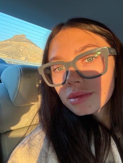 A young female is seated in the rear passenger seat of a vehicle, within the Arava Tichona Regional Council, Israel. She has dark hair, blue-green eyes, and wears oversized, thick-rimmed grey eyeglasses, with "GUCCI" embossed on the right temple. Her face is directly illuminated by strong, warm sunlight originating from the right, creating distinct highlights and shadows. Her lips are full. She is wearing a light-colored top. Through the vehicle's rear window, a large, light-colored natural rock formation with a pyramidal shape is visible against a clear blue sky, indicating an arid or desert environment.