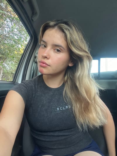The scene depicts a young female inside a vehicle. While the requested location is Santa Rosa, Argentina, no specific visual identifiers within the image confirm an exact street or area within the city. The individual, appearing to be in the passenger seat, has light skin, long wavy blonde hair with darker roots, and is looking directly at the camera. She wears a dark grey, ribbed short-sleeve top with faint white text on the chest, which includes "RCLMD" and "COLOMBIA". Dark-colored shorts are partially visible. A reddish mark is present on her chin below the lower lip, and a lighter mark above the upper lip. The interior of the vehicle features a light-colored roof lining and door frame. Through the open driver's side window, green foliage from trees is visible, indicating an outdoor setting. No other individuals or explicit interactions are discernible.