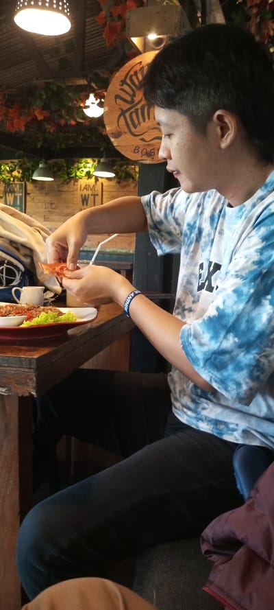 An Asian man wearing a blue tie-dye shirt and dark pants is seated at a wooden table. He is holding food in his hands and preparing to eat from a plate of rice and lettuce. A clear glass with a straw and a small cup sit on the table next to the plate. The restaurant has hanging plants, pendant lights, and a wooden sign. He is wearing a blue wristband.