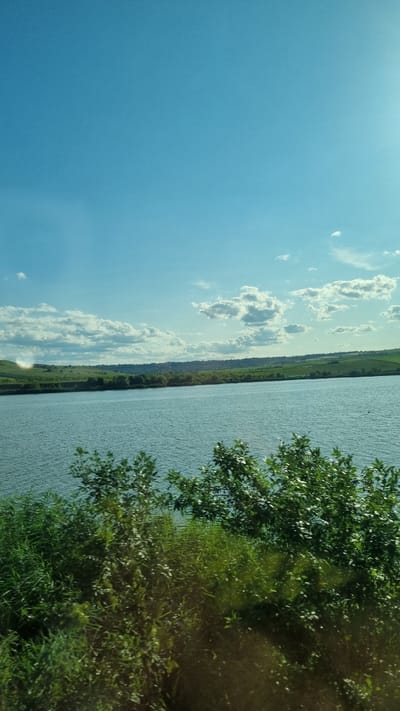 The image captures a scenic view of a body of water bordered by lush green hills under a bright blue sky dotted with fluffy white clouds. In the foreground, dense green foliage and trees frame the scene. The water appears calm with gentle ripples, reflecting the light. The landscape suggests a rural or natural setting, possibly a reservoir or a large lake. The hills in the background are covered in green grass and scattered with trees, with what appears to be a settlement or scattered buildings visible on the horizon. The overall impression is one of tranquility and natural beauty. This scene could be representative of the picturesque landscapes found in and around Pocoleni, Romania, known for its rolling hills and open spaces.
