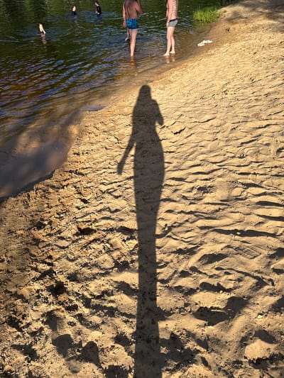 This is a high-angle, full shot of a sandy embankment next to a body of water. The image is oriented vertically, with the water on the left and the sand on the right.
**Subjects:**
On the left side of the image, partially submerged in the water, are three people. The person closest to the top of the image is lying down, with their legs visible above the water. This person is wearing blue patterned shorts and has tattoos on their legs. Below them, two other individuals are also in the water, appearing to be swimming or relaxing. On the sand, near the water's edge, a pair of white flip-flops are visible.
**Scene/Setting:**
The scene is a natural outdoor environment, likely a lake or riverbank. The water is dark and reflective, with ripples on the surface. The embankment is composed of light-colored sand, with numerous textured indentations and patterns, possibly from footprints or natural erosion. Shadows are cast across the sand, indicating a sunny day.
**Activity/Event:**
The people appear to be enjoying a day by the water, possibly swimming or wading. The presence of flip-flops suggests a casual recreational outing.
**Notable Details:**
The long, distinct shadow of a person stretches across the sand, suggesting the sun is low in the sky, likely late afternoon or early morning. The overall lighting and the warmth of the sand's color suggest pleasant weather.
**Text:**
There is no visible text in the image.
**Location Context:** Глівінскі сельскі Савет, Belarus