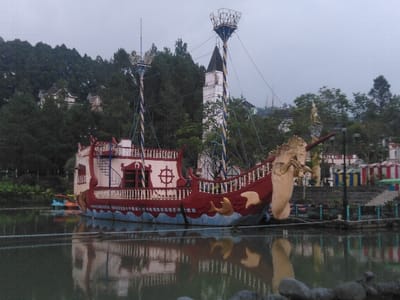 An ornate, stationary amusement park boat, designed to resemble a galleon, sits on a placid body of water under an overcast sky. The boat is predominantly red on its hull, featuring white railings and an upper deck structure. Its bow is adorned with a cream-colored unicorn figurehead, complete with a golden horn, and gold wave-like patterns and fish cutouts on the lower hull. A white spiraling staircase leads to an upper level, which also features a red door and a decorative white ship's wheel. A tall, striped mast (blue and white/yellow) rises from the boat, supporting a crow's nest-like structure at its apex. The still water reflects the boat's colors and form. In the background, to the left, a steep hillside is densely forested with dark green coniferous trees, among which several multi-story buildings with light walls and dark roofs are visible. Centered behind the boat is a white, steeple-like building with a dark, pointed roof, resembling a lighthouse. To the far right, additional trees and various colorful amusement park structures are discernible. No people are visible. The scene is located in Parque de Diversiones, Ciudad de San José, Costa Rica.