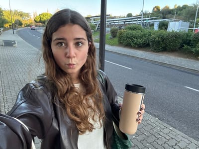 A young woman is prominently featured in a selfie-style photograph taken outdoors in Porto Salvo, Portugal. She has long, wavy brown hair, dark eyes, and her lips are puckered. She is wearing a dark brown jacket over a light-colored top. In her right hand, she holds a cylindrical beige and black thermos; her fingernails are painted with dark polish.

The immediate environment includes an interlocking gray paver sidewalk to her left and foreground. A dark asphalt road with a white dashed centerline curves into the distance. Two light-colored vehicles are visible on the road in the background. A dark street light pole is positioned behind the woman. Beyond the road and a strip of green shrubbery, a hillside displays multiple rows of ground-mounted solar panels. Trees populate the hillside and roadside. A concrete block marked "TIX" is partially visible on the sidewalk. The sky is clear blue.