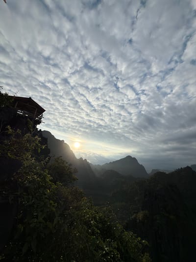 The image depicts a scenic overlook in Naka, Laos, featuring a rustic wooden structure perched on a rocky cliff edge. The structure appears to be an open-air shelter with a sloped roof, likely serving as a viewpoint or resting place. Lush green foliage, including trees and bushes, surrounds the shelter and drapes over the cliff, obscuring some of the rock face.

In the background, a dramatic mountain range unfolds, its peaks silhouetted against a sky filled with textured, wave-like clouds. The sun, a bright orb, is visible through a gap in the clouds, casting a warm glow and long shadows across the landscape, suggesting either sunrise or sunset. The light rays pierce through the clouds and illuminate the distant mountains, creating a sense of depth and atmosphere. The overall environment is natural and mountainous, with the human-made structure integrated into the rugged terrain.