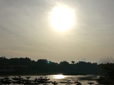 A wide shot captures a tranquil natural landscape with a body of water in the foreground, reflecting the bright, hazy sun. The sun is a prominent, glowing orb in the overcast sky. Silhouetted against the light are lush, dark green trees and hills that form the background. In the lower left, a dark-colored building with a sloped roof is partially visible. The water's surface is calm, with ripples creating shimmering patterns. The overall impression is one of peace and natural beauty, likely taken at dusk or dawn given the diffused sunlight and the silhouettes.