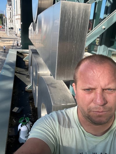 A man with fair skin, blue eyes, and short brown hair takes a selfie in front of a large, abstract, metallic sculpture in a city. He is wearing a light green t-shirt. The sculpture appears to be composed of large, curved metal shapes. In the background, to the left, is a street with a few people walking and one person on a bicycle. There are buildings and flags visible along the street. To the right of the sculpture, there is a modern-looking green and glass structure. In the lower left corner of the image, a white spray bottle with a green nozzle and handle is visible, resting on the ground next to some debris. The overall setting appears to be an outdoor public space in a city. The man's face is the most prominent subject in the foreground, with the sculpture and cityscape serving as the backdrop. The scene is lit by sunlight, casting shadows on the ground.
