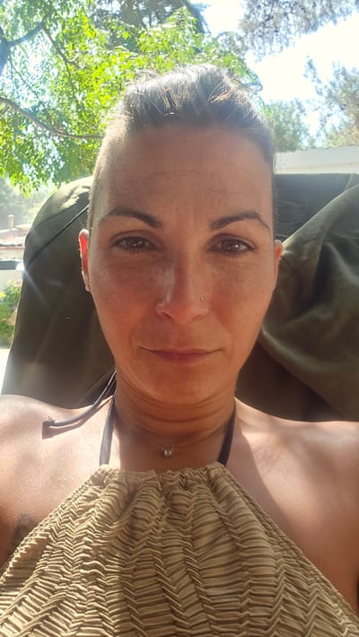 A woman is pictured in a selfie taken outdoors, likely in Toroni Municipal Unit, Greece, given the presence of trees and what appears to be sunlight filtering through foliage. She is seated, with the back of what seems to be a chair or lounge covered in a dark green fabric behind her. The woman has her hair pulled back and is wearing a necklace with a small square pendant. She is also wearing a textured, light brown halter-top. Her skin appears to be tanned, and she has a small nose piercing. The lighting suggests it is daytime and sunny, with some dappled sunlight visible on her face and shoulders.