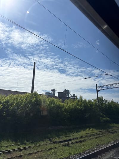 The image depicts an outdoor scene in Vyatskiye Polyany, Russia, viewed from a train window. The upper section features a bright blue sky with scattered white cumulus clouds, displaying a prominent sun flare in the upper left quadrant. Several dark electrical catenary wires traverse horizontally across the sky, with associated insulators and support structures visible. Below the sky, a dense line of vibrant green trees and bushes extends across the midground. Behind and above this vegetation, industrial structures are visible, including a tall, slender, dark cylindrical chimney on the left, and multiple metallic processing units or silos with platforms and piping in the center-right. A lattice-work railway gantry or support structure is also present on the far right. The foreground shows multiple parallel railway tracks. The tracks in the center-left are significantly overgrown with green grass and weeds, while the track closest to the viewer on the right has visible ballast and appears active. No individuals or animals are present in the scene. The overall lighting suggests bright daylight conditions.