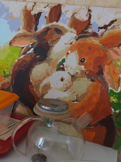 A painting of giraffes is displayed on a wall. In the foreground, a glass teapot with a silver lid sits on a white surface. To the left of the teapot is an orange object. The background is a light-colored wall. The image is taken inside a room in Shakhtarsk, Ukraine.