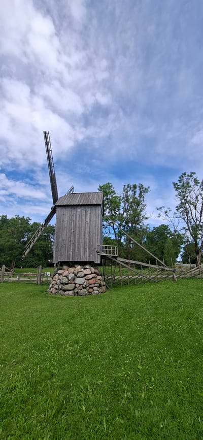 The image depicts a historical wooden windmill situated in a green, grassy field under a cloudy blue sky. The windmill is constructed of weathered grey wood and stands on a circular base made of large, rough stones. One of its sails is extended upwards at an angle, while the other is not fully visible. A wooden ramp leads to a small platform attached to the side of the windmill. A rustic wooden fence runs across the background, separating the field from a line of trees. The environment appears to be a rural or park-like setting, characteristic of a historical village or museum. This windmill is located in the village of Angla, on the island of Saaremaa, Estonia, known for its windmill park. There are no people visible in the image.