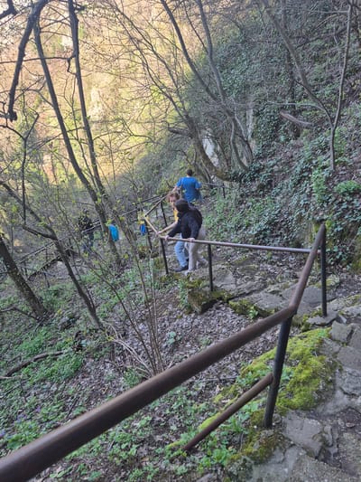 The image shows a downward view of a section of a stone staircase with a metal railing, located in a wooded area, likely a park or nature preserve. A woman wearing a black jacket and light-colored pants is walking down the stairs. Behind her is another person wearing a blue jacket and carrying a blue backpack. The surrounding environment includes trees with bare branches and some green vegetation on the ground and along the stone steps. Moss grows on the stone and railings. The location is Ivanovo, Bulgaria.