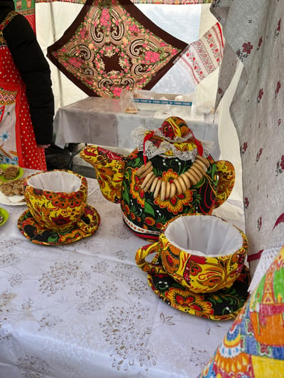 This image captures a vibrant display of folk art and traditional items, likely at a market, festival, or craft fair. The scene is illuminated by natural light, suggesting it is daytime, possibly outdoors or under a large canopy.
In the foreground, slightly to the right, are two fabric cozies. The most prominent is a large teapot cozy, intricately decorated with a rich, colorful floral pattern featuring yellow, red, orange, and green flowers and leaves on a black background, reminiscent of Khokhloma painting. A string of light-colored, ring-shaped bagels (known as sushki or baranki, traditional Eastern European snacks) is draped over the top of the teapot cozy. Next to it, to the lower right, is a smaller, matching cozy, possibly for a cup or a smaller pot, with the same vibrant floral design and a white lining visible inside. These cozies rest on a light-colored tablecloth or fabric which has a subtle, almost golden, botanical pattern.
To the left, a dark brown square scarf or shawl is spread out, displaying a large, elaborate pattern of pink, cream, and brown paisley motifs and flowers. Above this scarf, another piece of white fabric with red embroidered patterns is visible.
In the lower left corner, an arm wearing a black sleeve is partially visible, extending towards a red fabric with white polka dots. Small, light green items, possibly slippers or decorative mats, are visible at the very bottom right of the image.
The background shows elements of a temporary structure, such as a tent or stall. There are various other fabrics and textiles in the background, including a colorful one with bright orange, yellow, and blue geometric or folk patterns. On a blue and white strip of material visible in the mid-background, partially obscured, the numbers "1400-1500" and "1500-1600" are discernible, which could indicate prices, size ranges, or time slots. The overall impression is one of cultural celebration and the showcasing of handcrafted goods.
