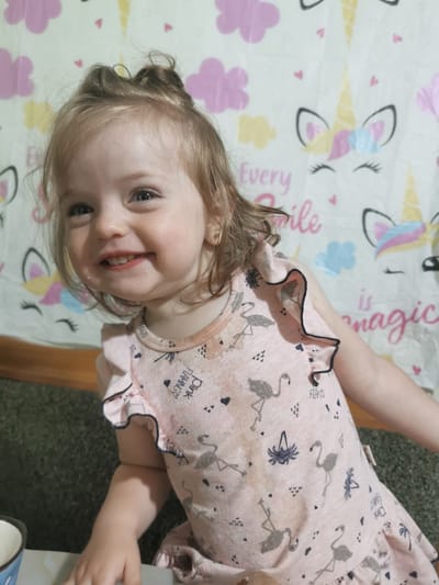 In Carbuna, Moldova, a young Caucasian girl with blonde hair is captured from the chest up, smiling. She is wearing a light pink dress adorned with a pattern of flamingos and the words "Pink Flamingo." The background is a light-colored fabric featuring a repeating pattern of unicorn faces, clouds, and the words "Every smile is magic." The lighting appears to be from a flash.