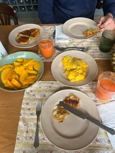 A top-down, medium shot shows a breakfast scene on a wooden table covered with a floral tablecloth. A person wearing a navy blue long-sleeved shirt holds a tan plate with a piece of croissant on it. Another tan plate contains scrambled eggs with black pepper. A third tan plate holds two pieces of waffles with a silver knife resting on it. A teal bowl filled with sliced mangoes is also present. Two glasses of orange juice are visible, along with a green patterned glass and a bowl of olives. A fourth tan plate holds two grilled cheese sandwiches. The scene appears to be set in a residential area, but there are no clear indications that place the scene in Gondarém, Portugal.