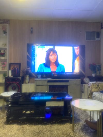 The image depicts an indoor residential living space in Akpajo, Nigeria. A large flat-screen television is centrally positioned against a light beige wall featuring vertical paneling. The TV screen displays a woman with dark hair, wearing a turquoise blazer, from a program identified as "SHOWCASE."

Below the television, a multi-component entertainment unit is present. To the left of the TV stand, a large black speaker system with red accents is visible, topped with a white air freshener. Adjacent to the speaker, a framed photograph of a person in academic regalia is displayed. The TV stand itself consists of light-colored cabinets with drawers. A light-colored vase holding dried flowers is on the left section of the TV stand. To the right of the TV, a small pink analog clock with white numbers sits on a surface.

To the far left, a tall, narrow white shelving unit displays decorative items, including pink artificial flowers in various vases and a small green plant. A golden decorative object is also on one of the shelves. A dark, possibly stylized bow tie-shaped wall decoration is mounted on the textured wall above the TV's left side. In the upper right corner of the room, a window-mounted or wall-mounted air conditioning unit is visible.

A black, ornate coffee table with gold trim is centered on a light-colored, shaggy rug covering the floor. The coffee table's surface holds several remote controls, some emitting a blue light. A lower shelf of the coffee table contains a basket, also with a blue glow. Two small, round or oval side tables with white marble-patterned tops and golden metal legs are positioned to the right of the coffee table. Part of a light-colored, patterned sofa or armchair with gold accents is visible on the far right, and a human foot is partially visible near the rightmost side table, suggesting an occupant. A small potted plant with red roses and a framed picture are on a surface next to the sofa on the right. The ceiling is white and paneled.