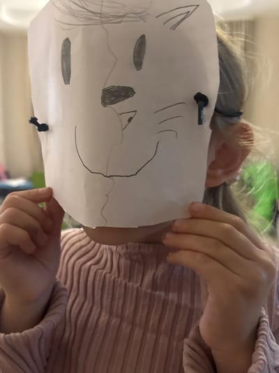 A child, whose face is mostly obscured by a homemade paper mask, is the primary subject of this portrait. The mask is white and appears to have been drawn on with charcoal or a dark pencil, depicting a stylized animal face with prominent ears and eyes. The child's fair hair is pulled back with a dark elastic band. They are wearing a light pink, ribbed shirt.

The child is holding the mask up with their hands, suggesting they are either showing it off or about to put it on. One hand is visible on the right side of the mask, while the other is partially obscured on the left. The background is blurred, but appears to be an indoor setting, with hints of green and possibly furniture. The lighting suggests daytime, and there are no obvious cues about the weather. The overall mood seems playful and creative, as the child is engaged with their craft. There is no visible text in the image.