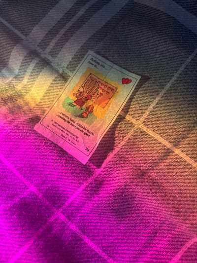 A close-up shot captures a "Love Is..." comic strip card lying on a textured surface, possibly fabric like a blanket or a towel. The card depicts two cartoon characters, a woman and a child, surrounded by hearts. The text on the card is in both Russian and English, with the Russian phrase "Любовь это..." and English "Love Is..." above the illustration. Below the image, there are additional lines of text in both languages: "...считать часы, когда вы врозь." and "...counting the hours you are apart." The bottom of the card features copyright information: "© 1997 MINIKIM CARIBBEAN NV. Kent Gıda Mad. San. ve Tic. A.Ş." and "Серия 7, 7th Series", along with the number "79".
The scene is illuminated by a gradient of light, transitioning from a warm yellow-orange on the left to a vibrant pink-purple on the right, creating an abstract and moody atmosphere. This lighting suggests either artificial lighting with colored gels or a unique photographic effect. The background surface has a visible weave pattern, with diagonal lines suggesting a plaid or checkered design. The overall impression is intimate and perhaps a bit melancholic, emphasized by the theme of distance in the comic and the dramatic lighting. No specific time of day, weather, or explicit location cues are visible beyond the generic surface and lighting. The context provided by the user of "Velykyi Sukhodil, Ukraine" is external information not discernible from the image itself.