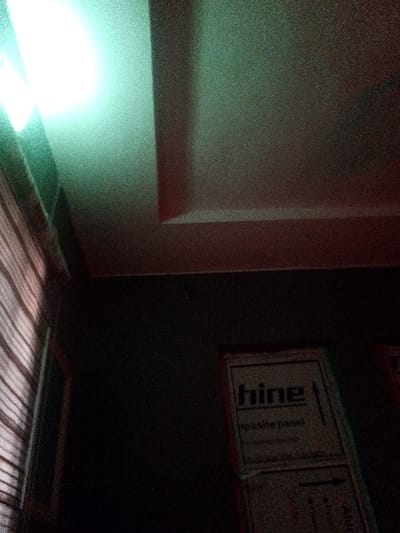 The image displays an interior scene in Kongo, Nigeria, characterized by low light and pronounced image noise. A bright, overexposed greenish-white light source is visible in the upper left quadrant, illuminating a portion of the light-colored, stepped tray ceiling. The ceiling exhibits a recessed architectural design towards the upper right. The lower two-thirds of the scene are dominated by a dark wall. On the far left, a window covered by light-toned horizontal blinds is partially visible. In the lower right, two rectangular objects, appearing to be packaged composite panels, lean vertically against the dark wall. The upper panel prominently displays the brand name "hine" with an upward arrow graphic, and the text "composite panel" beneath it, rendered in dark lettering on a lighter surface with a reddish-brown border. No individuals, actions, or direct interactions are discernible within the frame.