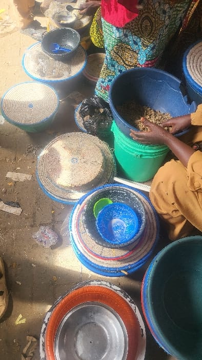 The scene depicts an outdoor or semi-outdoor work/market area in Fagge D1, Nigeria, under direct sunlight, evidenced by harsh shadows. The ground is light brown dirt with scattered debris.
Multiple circular woven baskets and plastic containers are arranged on the ground. Notable objects include:
- A large blue plastic basin containing a light brown granular substance, possibly grain or seeds, resting on a green plastic bucket. The hands and lower torso, clad in a light brown garment, of an individual are visible, actively manipulating the substance within the blue basin.
- Several shallow, circular woven baskets of various sizes, many with blue-painted rims or bases, are present. One holds a black plastic bowl with a blue plastic scoop. Another basket supports a stack of smaller blue and green plastic bowls.
- A metallic, circular basin with an orange rubber rim is positioned in the lower-center.
- A partially visible individual in the upper-center wears a vibrant patterned fabric (blue, green, yellow, red motifs) with a red top.
- A light-colored sandal, belonging to another individual, is visible in the lower-left.
- Black plastic bags containing dark solid material are seen on some baskets.