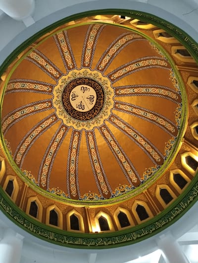 The image displays an ornate dome ceiling, likely within a mosque, featuring intricate Islamic calligraphy and geometric patterns. The central part of the dome is adorned with Arabic script arranged in a circular pattern, surrounded by radial bands of decorative motifs. These bands are composed of interlocking geometric shapes and stylized floral elements in colors such as gold, brown, white, and muted blues and reds. The dome's exterior edge is bordered by a green band featuring more elaborate calligraphy. Below this, a series of arched windows with golden frames are visible, set against a brown, textured background. The overall impression is one of rich architectural detail and artistic craftsmanship. The image appears to be taken from below, looking upwards towards the apex of the dome. The location is identified as the city of Blangmetimu, Indonesia.