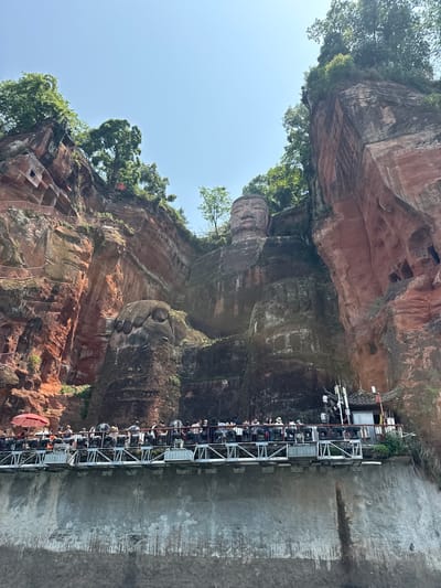 The image features a monumental rock carving in Shizhong, China, likely the Leshan Giant Buddha. The figure is sculpted directly into a cliff face, with red sandstone visible. A metal walkway with multiple levels has been constructed to allow visitors to view the sculpture. People are visible walking on the walkway, observing the carving. Trees and other vegetation grow atop the cliff face. The sky is light blue.