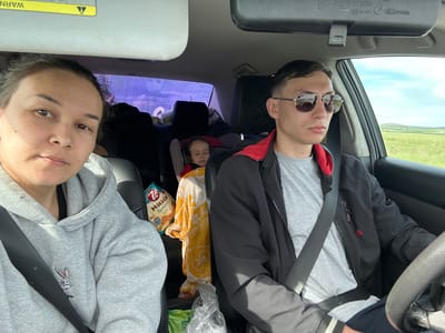 The image depicts the interior of a car during daytime, occupied by three individuals: two adults in the front seats and one child in the back.

On the left, a woman, appearing to be the passenger, is taking the photograph. She has dark hair, a neutral expression, and is wearing a light grey hooded sweatshirt with an embroidered white Bugs Bunny character on the left chest. A black seatbelt crosses her torso. Above her, a sun visor is visible, showing an inverted yellow warning label with black text and an exclamation mark symbol. To her right, a package of "7 дней МИНИ Круассаны" (7 days MINI Croissants) is visible on the seat or console.

On the right, a man is driving the vehicle. He has short dark hair, is wearing aviator-style sunglasses that reflect the outside landscape, a grey T-shirt, and a black zippered jacket with red lining visible at the collar. A small red Reebok logo is present on the right side of his jacket. A black seatbelt is fastened across his chest, and his right hand is partially visible on the steering wheel. Above him, a rearview mirror shows markings, including "01 5709."

In the center of the back seat, a small child with dark curly hair is visible, appearing to be asleep in a car seat. The child is partially covered by a yellow blanket or garment with a white patterned design.

Through the windows, particularly the passenger-side front window, a vast rural landscape is visible, characterized by green fields, gently rolling hills in the distance, and a light blue sky with scattered white clouds. The scene suggests the car is traveling through an open area near what is identified as Sukhtelinskiy, Russia. Inside the car, clear plastic bags and a green bag are visible on the floor or between the seats.