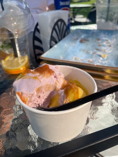 In Znamianka, Ukraine, a close-up shot captures a white paper cup filled with two scoops of ice cream, one pink and one yellow, topped with a caramel sauce. A black spoon rests in the cup. In the background, a clear plastic cup with a straw, lemon slice, and mint sprigs is visible, along with a stack of blue and white plates with remnants of food, and a napkin dispenser. The scene is set on a glass table, with hints of greenery and outdoor elements suggesting a cafe or restaurant setting.