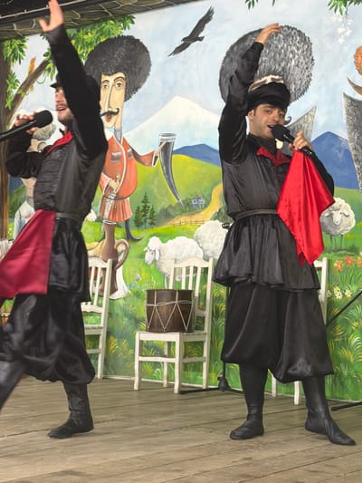 In Kveda Chkhutuneti, Georgia, two male performers in traditional black and red costumes are on a wooden stage in front of a colorful mural depicting a pastoral scene. The performer on the left, with a raised left arm, holds a microphone in his right hand and wears a black hat and a red sash. The performer on the right, also with a raised arm, holds a microphone in his left hand and a red cloth in his right, wearing a similar black hat with a feathered adornment. The mural features stylized human figures, sheep, trees, mountains, and a bird in flight. Two white chairs flank the performers, and a drum sits on a platform behind the performer on the right.