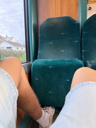 The image shows a close-up view from the perspective of someone sitting on a train. The train seats are upholstered in dark teal fabric with a repeating pattern of small, abstract arrow shapes in yellow and blue. The person's bare legs are visible, wearing light blue denim shorts and white socks with athletic shoes. Outside the window, there are houses and trees, suggesting a suburban or semi-rural setting. The sky is overcast. The interior of the train car features wood paneling on the walls and a green frame around the window. There is a vent visible below the window. The overall impression is of a person traveling by train.