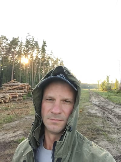 A man wearing a hooded jacket and a baseball cap is taking a selfie in a forest. The sun is setting behind the trees, casting a warm glow on the scene. There is a pile of cut logs to the left of the man, and a dirt road stretches into the distance to his right. The environment appears to be a logging area in the village of Tsyarekhovichy, Belarus.
