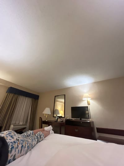 A person is lying in bed wearing white socks and floral pajama pants. A TV and a vanity with a mirror and lamps are visible. A window with striped curtains is present. The location appears to be a hotel room in San Nicolás de los Garza, Mexico.