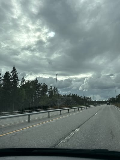 The image depicts a road scene in Ørje, Norway, characterized by an overcast sky filled with dramatic, grey clouds. A bright patch of sunlight breaks through the clouds in the upper left quadrant. The road, a two-lane asphalt highway, stretches into the distance, bordered by a metal guardrail on the left and a dense forest of evergreen trees on the right. The trees are tall and dark, with some rocks visible among them on the left side of the road. Multiple streetlights are spaced along the right side of the highway. A dark-colored car is visible in the distance on the right lane. The image appears to have been taken from the perspective of someone inside a vehicle, indicated by the black dashboard visible at the bottom of the frame.