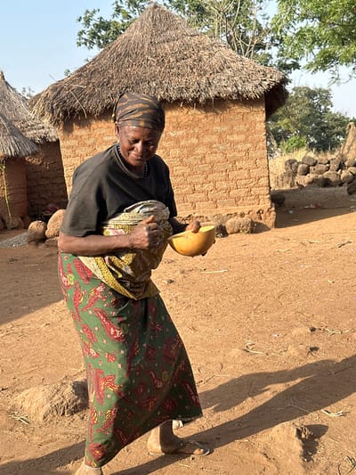 An elderly woman stands in a rural setting, possibly a village, near a thatched-roof building with mud brick walls. She wears a dark top, a patterned green and red skirt, and a headscarf. Her hands hold a yellow gourd and some other cloth materials. The ground is dry and dusty, with a stark shadow visible from the sunlight. The environment appears to be warm, with sparse vegetation.