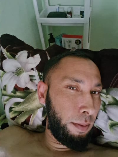 The image is a selfie taken from a low angle, focusing on a man lying down. He has a beard and is looking directly at the camera. The man is resting on a pillow with a floral pattern, featuring white lilies and green leaves. Behind him, a white shelf unit is visible, holding several bottles and a red and white box. The wall behind the shelf is a pale green. The overall setting appears to be indoors, possibly a bedroom, given the pillow and shelf. There is no explicit indication of the city of Kahramon, Uzbekistan, within the image itself.