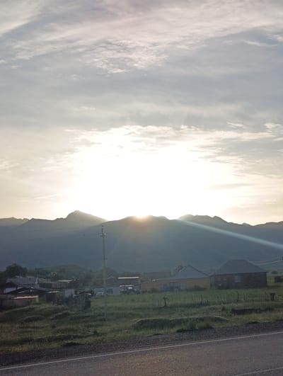 The image shows a rural landscape at sunrise in Kyzyl-Jyldyz, Kyrgyzstan. In the foreground, a paved road is visible, with a grassy verge separating it from a field. Several buildings, including houses and what appears to be a barn, are scattered across the field. A tractor is parked near one of the buildings. In the middle ground, a power line pole stands prominently. The background features a silhouette of mountains against a bright, sunlit sky with scattered clouds. The sun is low on the horizon, casting rays of light through the clouds and over the mountains. The overall impression is of a peaceful morning in a rural setting.