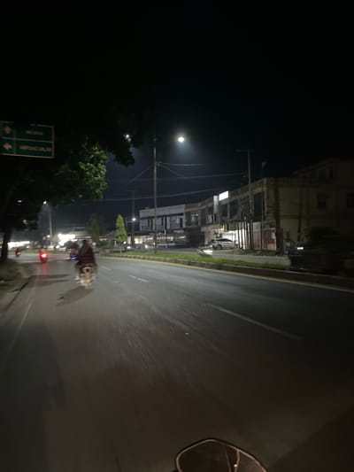 The image depicts a street scene at night in a location that appears to be in Indonesia, specifically Dalam, based on the provided context. The scene is illuminated by streetlights and the moon, creating a dark yet visible environment.

The main subjects are a street, buildings, trees, and a few vehicles, including a motorcycle with a rider. The street is wet, suggesting recent rain or that it is actively raining, and the surface reflects the available light. The buildings lining the street appear to be commercial or residential, with some windows lit. Lush green trees are visible on the left side of the frame, contrasting with the urban surroundings.

A motorcycle rider, holding an umbrella, is a prominent element in the foreground, indicating that the rider is likely using the umbrella to shield themselves from the rain while riding. Other vehicles, including what looks like a car and possibly other motorcycles, can be seen further down the street, their lights adding to the illumination.

The time of day is clearly night, characterized by the dark sky and artificial lighting. The presence of rain is a notable detail that contributes to the mood of the scene, which is somewhat solitary and perhaps even melancholic due to the darkness and the wet conditions. The overall atmosphere is that of a quiet, wet night on a street with some activity.

There is visible text on a green street sign on the left side of the image. The text is in Indonesian, and while partially obscured, it appears to include directional arrows and possibly place names.