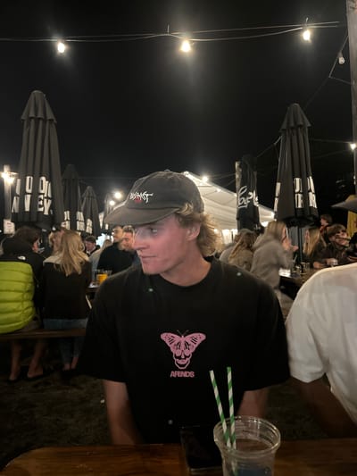 The image depicts an outdoor, nighttime social gathering at a venue in Wombarra, Australia. The foreground is dominated by a male individual, light-skinned, with wavy blonde hair, wearing a dark baseball cap featuring white script and a black t-shirt with a pink butterfly and skull graphic and "AFENDS" text. He is seated at a wooden table, looking towards his left (viewer's right). On the table in front of him are a clear plastic cup containing a blue liquid with two green and white striped straws, and a dark smartphone.

To the immediate right of the primary subject, a portion of another person in a white t-shirt is visible. To the primary subject's left, a person wearing a bright yellow-green puffer jacket is seated at the same or an adjacent wooden table. The midground and background contain approximately 15-20 additional individuals, mostly seated at similar wooden tables and benches, engaged in various states of socialization. Several large, dark-colored, closed patio umbrellas are spaced throughout the area, some displaying partial white text. Overhead, strings of incandescent lights illuminate the scene against a dark night sky. The ground surface appears to be dark, uneven earth or gravel. A white tent or temporary structure is visible in the distant background. No specific direct interactions are discernable from the primary subject.