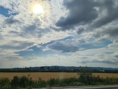 A wide-angle shot captures a rural landscape under a cloudy sky with the sun partially obscured by clouds. In the foreground, a patch of green bushes and foliage lines a grey surface, possibly a road or path. Beyond this, a vast, dry, golden-brown field stretches across the middle ground. A line of trees and shrubs forms a dark green band separating the field from the background. In the far distance, a range of blue, undulating hills or mountains forms the horizon. The sky is a dynamic mix of bright white and grey clouds, with shafts of sunlight piercing through in the upper left quadrant. A hint of blue sky is visible between the clouds. On the horizon, a few structures, possibly buildings or infrastructure, are visible in the distance, as are a few vehicles including a white van and a red truck with a trailer. The image appears to be taken from a moving vehicle, given the slight blur and the perspective. The overall scene is serene and depicts a countryside setting. The location is identified as Wiener Neustadt, Austria.