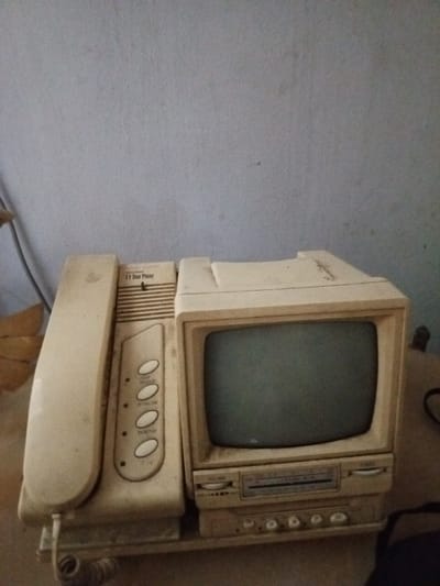 The image displays a stationary, cream-colored vintage video door phone system resting on a dusty, light-colored flat surface against a plain, muted light blue-grey wall in Keo Kouang, Laos. No individuals are present.
The system comprises three integrated components. On the left, a handheld telephone receiver, connected by a coiled cord, rests off its cradle. Adjacent to it, a central control panel features vertical ventilation grilles at its top, with the label "TV Door Phone" inscribed below. Below this are four prominent oval buttons, labeled from top to bottom: "TALK / RELEASE", "ROOM INTERCOM", "DOOR OPEN", and "MONITOR", followed by a smaller circular button. To the right, an integrated small Cathode Ray Tube (CRT) monitor is visible, displaying a dark, inactive screen. Beneath the monitor, a horizontal control strip includes a linear dial and two rotary knobs, one explicitly labeled "VOLUME". Below this strip, a lower panel contains four smaller circular buttons or knobs.
The entire unit shows extensive dust accumulation and yellowing discoloration, indicative of age and prolonged disuse. The background wall exhibits minor smudges and textural imperfections. A dark, coiled cable is partially visible on the far right of the frame, extending off-screen.