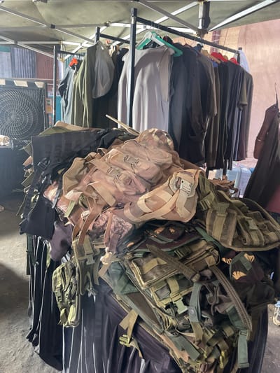 The image shows a stall under a pop-up canopy with clothing hanging on a rack on the left and a pile of military-style tactical vests and pouches on the right. The clothing rack is filled with garments in various shades of gray, brown, and black. The pile of tactical gear is in various colors including tan, olive green, and camouflage patterns. An electric fan is visible beneath the stall. The location is likely in Mira-Bhayander, India.